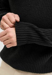 Czarny sweter dzianinowy o ribbowanej fakturze i dopasowanych mankietach. Zbliżenie na dłonie trzymające materiał, podkreślające szczegóły wykończenia i designu.