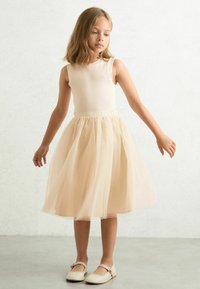 Bež majica bez rukava s višeslojnim, suknjom od šampanjac tulle do koljena. Suknja ima suptilan sjaj. U kombinaciji s svijetlim ravnim cipelama.