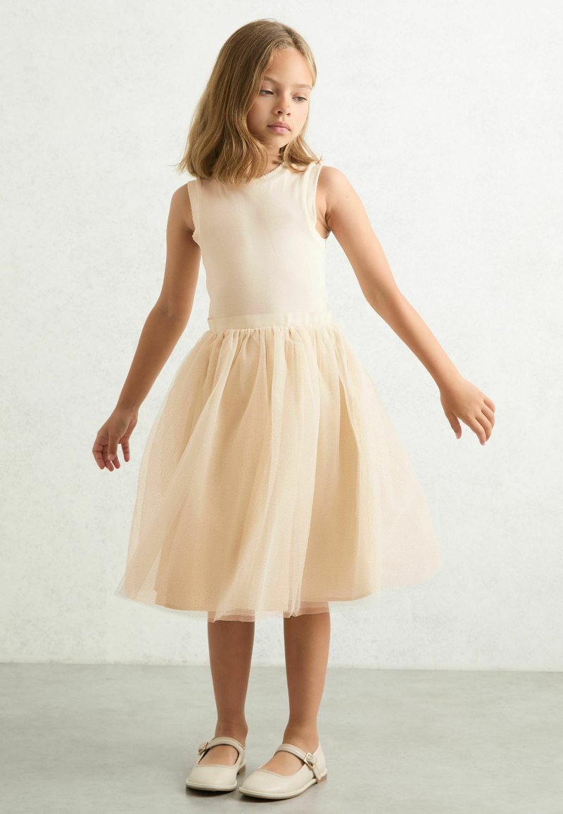 Bež majica bez rukava s višeslojnim, suknjom od šampanjac tulle do koljena. Suknja ima suptilan sjaj. U kombinaciji s svijetlim ravnim cipelama.