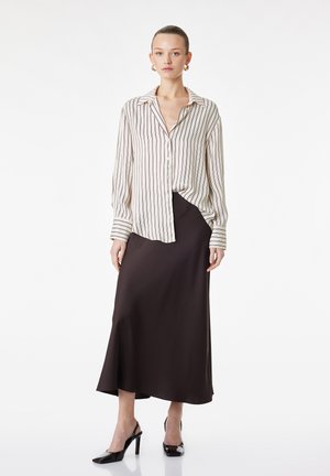 Frau trägt eine cremefarbene und braune, vertikal gestreifte Bluse, einen dunkelbraunen Midirock, schwarze hochhackige Slingback-Schuhe und goldene Creolen.