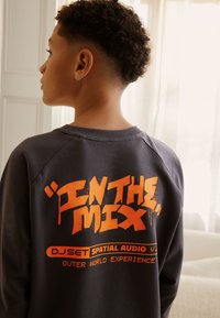 Grauer Sweatshirt mit großem orangefarbenem Grafikdesign auf der Rückseite und dem Text "IN THE MIX" sowie weiteren Markendetails.