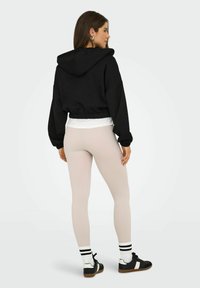 Haut de survêtement noir coupé avec capuche, ourlet élastique, associé à un legging côtelé rose clair et des baskets noires avec accents blancs et des chaussettes rayées.