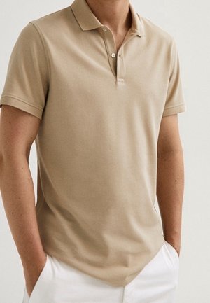Polo beige a maniche corte con colletto e due bottoni indossata da una persona con le mani nelle tasche dei pantaloni bianchi.
