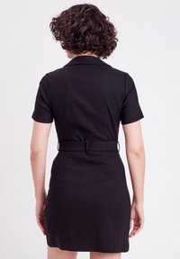 Femme aux cheveux bouclés portant une robe noire ceinturée à manches courtes, vue de dos sur un fond blanc uni.