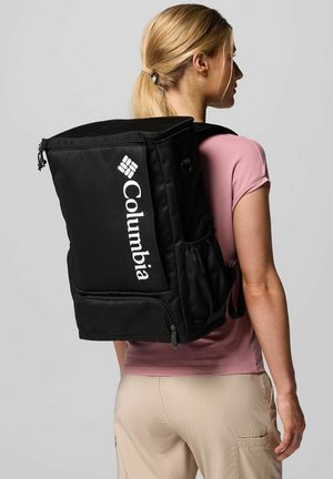 Sac à dos Columbia noir de forme rectangulaire, avec une poche zippée à l'avant et un logo blanc. Tissu texturé et bretelles réglables.