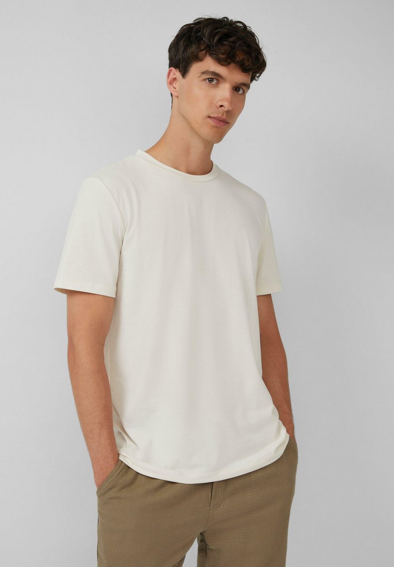 Koszulka w kolorze kremowym z krótkim rękawem i okrągłym dekoltem, wykonana z gładkiego materiału, w zestawieniu z teksturowanymi brązowymi spodniami. Prosty i neutralny design.