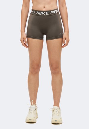 Un mannequin se tient devant un fond uni, portant un short ajusté Nike Pro foncé et des baskets Nike crème.