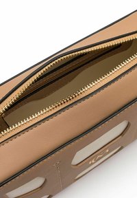 Sac en cuir marron avec une fermeture éclair dorée, présentant une texture lisse et des éléments de design découpés. L'intérieur est doublé d'un tissu clair.