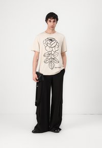 Beige bomulls t-skjorte med svart blomstermotiv, paret med løse svarte bukser og en liten crossbody-veske med stropp.