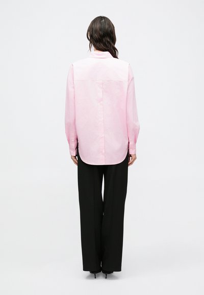 Ružová košeľa na gombíky s dlhými rukávmi, so zaobleným lemom a mierne voľným strihom, nosená s čiernymi širokými nohavicami.