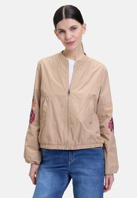 Beige bomberjack met een rits aan de voorkant, elastische manchetten, en geborduurde bloemenaccenten in paars en oranje op de mouwen.