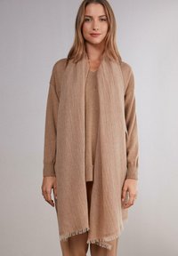Sciarpa beige realizzata in materiale morbido, con bordo frangiato e finitura tessurizzata, drappeggiata elegantemente su un maglione coordinato.