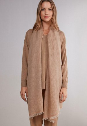Beige tørklæde lavet af blødt materiale med frynser og en tekstureret overflade, elegant draperet over en matchende sweater.