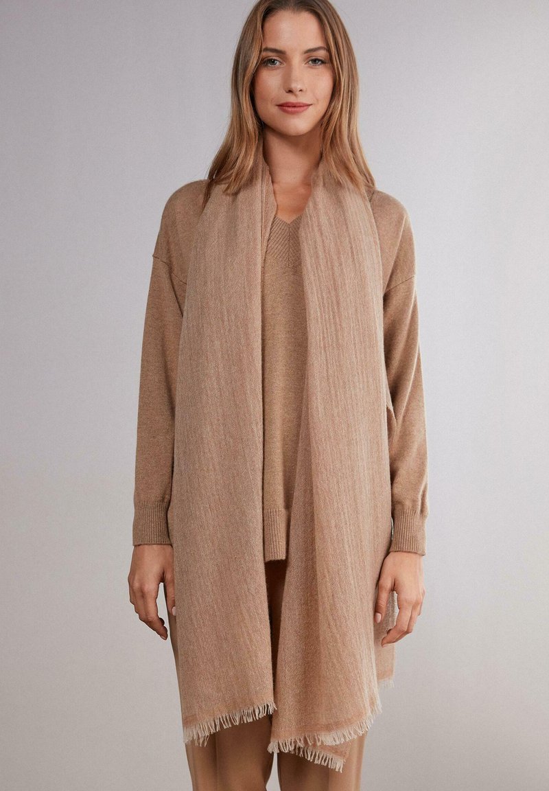Sciarpa beige realizzata in materiale morbido, con bordo frangiato e finitura tessurizzata, drappeggiata elegantemente su un maglione coordinato.