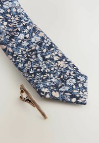 Cravate florale marine avec des motifs bleu clair et rose, fabriquée à partir d'un tissu texturé, accompagnée d'une pince à cravate en bronze avec un design en maille.