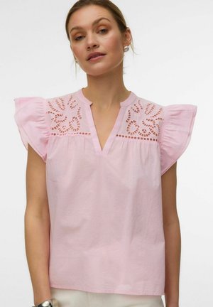 Femme portant un chemisier rose clair avec des détails découpés floraux aux épaules et des manches courtes à volants, debout devant un fond clair uni.