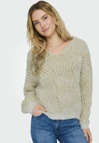 Cremefarbener, strukturierter Pullover mit V-Ausschnitt und gewelltem Muster. Hat lange Ärmel und eine lässige Passform, kombiniert mit blauen Jeans.