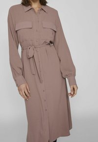 Dlouhé šaty s knoflíky v odstínu mauve. Mají dvě náprsní kapsy, pás se sponou a dlouhé rukávy s knoflíkovými manžetami. Měkká textura.