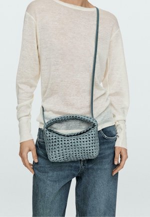 Person trägt ein hellbeiges langärmliges Oberteil und blaue Jeans, trägt eine kleine graue gewebte Umhängetasche mit langem Riemen.