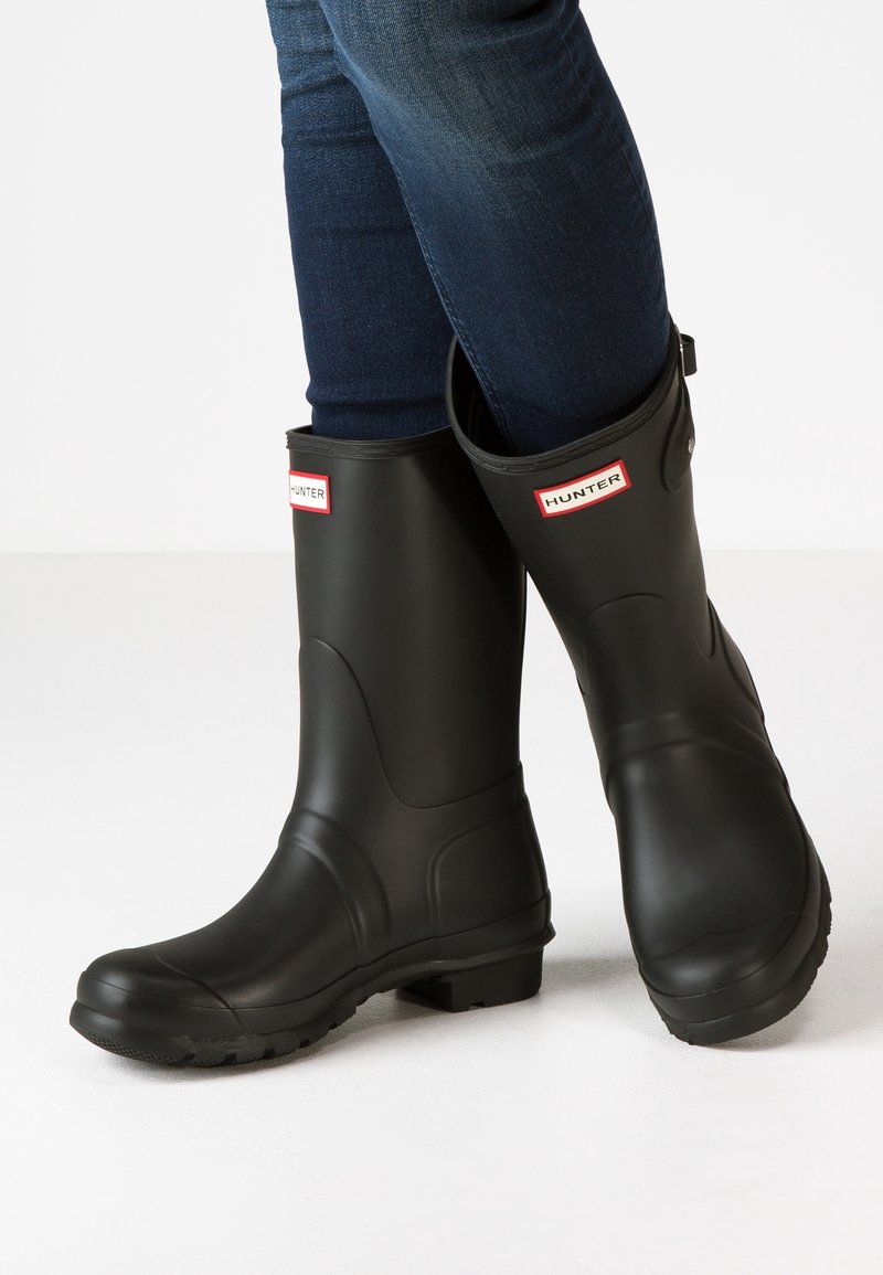 Osoba nosząca czarne kalosze Hunter z widocznymi dżinsami, stojąca z jedną stopą skrzyżowaną nad drugą na białej podłodze.