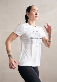 Valge sportlik t-särgis, halli tekstiga "ÄRA KUNAGI PEA STOPPI KONKUREERIMIST," lühikesed varrukad, sobiv disain, koos mustade retuusidega, sile tekstuur.