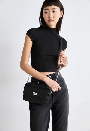 Jeune femme portant un crop top noir ajusté et un jean foncé, tenant un sac à épaule noir avec un logo argenté et une bandoulière chaîne.