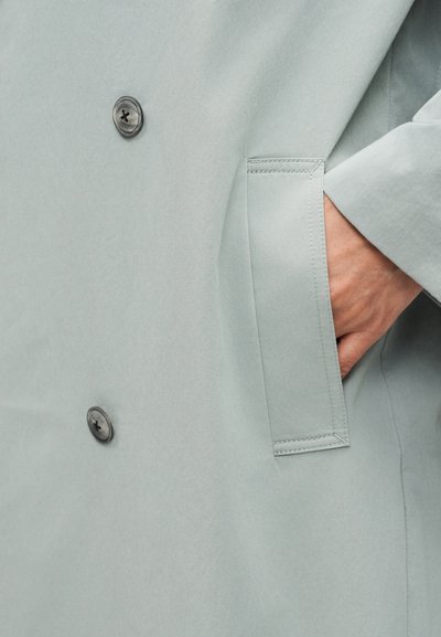 Χέρι βουτηγμένο στην πλαϊνή τσέπη μιας ανοιχτόχρωμης γκρι παλτό με ορατά κουμπιά και λεπτομερές ράψιμο στην άκρη της τσέπης.