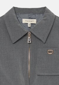 Sivi zip-up majica s oštrim ovratnikom, džepom na prsima s malim zlatnim grbom i etiketom "Twinset" unutar ovratnika.
