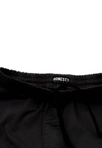 Schwarze Shorts mit einem elastischen Bund und Kordelzug. Verfügt über ein kleines weißes Etikett mit der Aufschrift "EHRLICHKEIT". Glatte Textur.