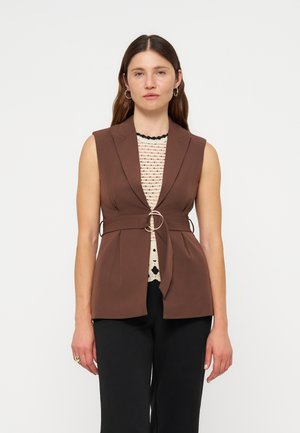 Kvinde, der kigger fremad, bærer en brun ærmeløs blazer med bælte, cremefarvet hæklet top, sorte bukser, store øreringe og har løsthængende brunt hår.