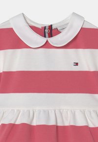 Růžové a bílé pruhované šaty s bílým límečkem ve stylu Peter Pan a malým vyšitým logem vlajky na hrudi.
