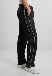 Pantaloni atletici negri cu o bandă albă pe lateral, talie elastică și buzunare frontale. Fabricat dintr-un material neted și ușor.