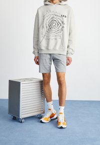 Světle šedá mikina s grafickým spirálovým vzorem, v kombinaci s šedými šortkami. Bílé a oranžové sportovní boty, bílé ponožky, stojící vedle kovového úložného kontejneru.