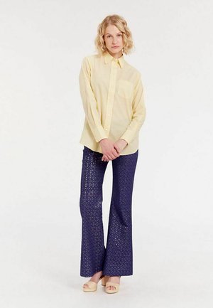 Chemise boutonnière jaune clair avec une poche, associée à un pantalon évasé bleu marine avec un motif en dentelle. Des chaussures compensées beiges complètent le look.