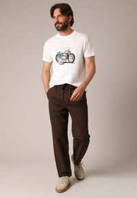 Witte katoenen t-shirt met motorfietsgraphic, gecombineerd met bruine broek en beige sneakers. Model staat casual met handen in de zakken.