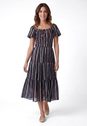 Lächelnde Frau, die ein schwarz-weiß gestreiftes Midikleid mit Blumenmuster, Puffärmeln und beigen Ballerinas trägt, vor weißem Hintergrund steht.