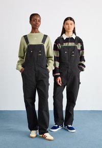 Zwarte overalls met voorzakken en verstelbare bandjes, gecombineerd met gelaagde sweatshirts. Schoeisel bestaat uit witte sneakers en blauwe schoenen.