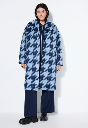 Kvinde iført en lang blå og marineblå hundetandsmønstret frakke over marineblå bukser og sorte robuste støvler, stående mod en hvid baggrund.