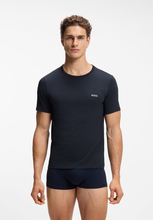 Giovane uomo con capelli corti e ricci che indossa una T-shirt nera aderente BOSS e boxer neri coordinati, in piedi davanti a uno sfondo bianco uniforme.