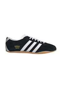 Melna zamšādas kedas ar baltām ādas detaļām, kurām sānos ir trīs svītras, gumijas zole un zeltīts "adidas TOKYO" logotips.