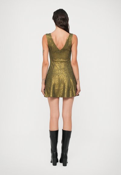 Auksinė palta su figūrą pabrėžiančiu siluetu ir plačia sijonine dalimi. Turi gilią V formos iškirptę nugaroje ir be rankovių dizainą. Su juodais iki kelių batais.