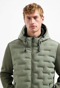 Jonge man met kort blond haar, gekleed in een groene gewatteerde hooded jacket met gestructureerde mouwen, recht vooruit kijkend tegen een witte achtergrond.