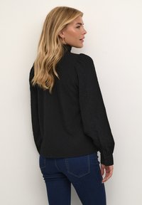 Blouse noire texturée à manches longues, col montant et poignets boutonnés, associée à un jean bleu ajusté. Design simple et élégant.