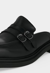 Μαύρο δερμάτινο παπούτσι χωρίς κορδόνια με διπλούς ασημένιους ιμάντες με αγκράφες και παχιά σόλα, απεικονισμένο σε απλό ανοιχτό φόντο.