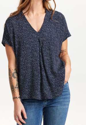 Femme portant un chemisier bleu marine à col en V avec des taches blanches, associé à un jean bleu ; des tatouages visibles sur l'avant-bras gauche et l'intérieur du bras droit.