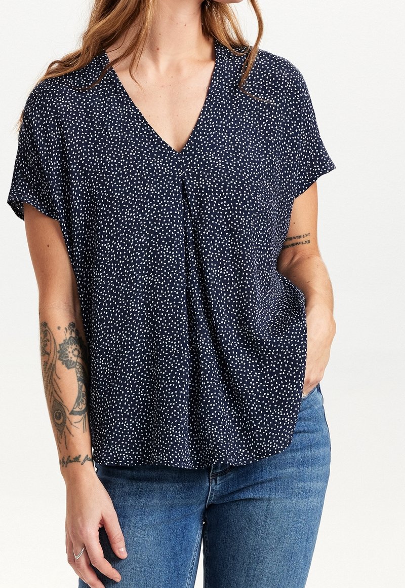 Femme portant un chemisier bleu marine à col en V avec des taches blanches, associé à un jean bleu ; des tatouages visibles sur l'avant-bras gauche et l'intérieur du bras droit.