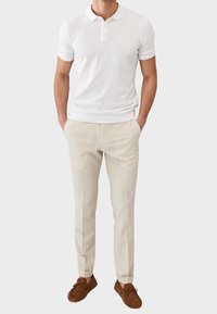 Camisa polo blanca de manga corta con cuello, combinada con pantalones cortos de un beige claro y zapatos de ante marrón con cordones. Textura de tela suave.
