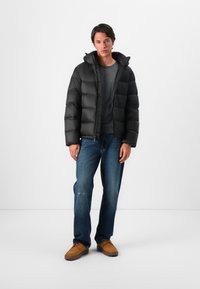 Zwarte pufferjack met een capuchon, gevoerd ontwerp en een voorkeursrits. Gecombineerd met een grijs t-shirt, blauwe jeans en een paar bruine schoenen.