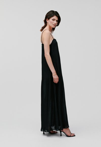 Black pleated maxi dress with thin straps, featuring a flowing, textured design and a slightly flared hemline, paired with strappy high heels.