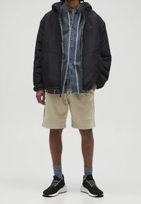 Czarna kurtka puchowa, w paski koszula zapinana na guziki w niebiesko-zielonym kolorze, beżowe szorty, czarne trampki z białymi podeszwami, szare skarpety, w pozycji stojącej.